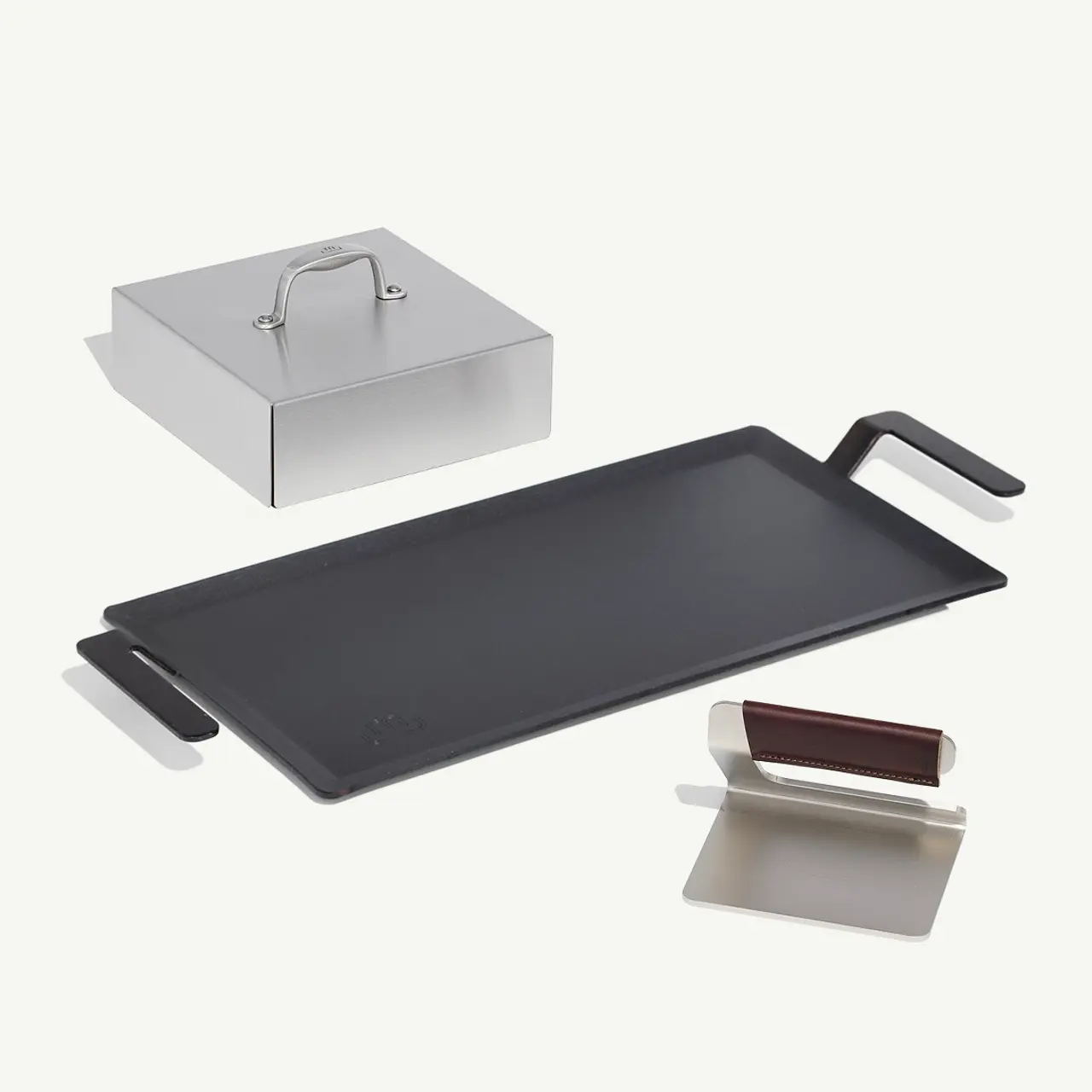 A flat, rectangular griddle with handles lies next to a small gray box with a handle and two metal spatulas on a light background.