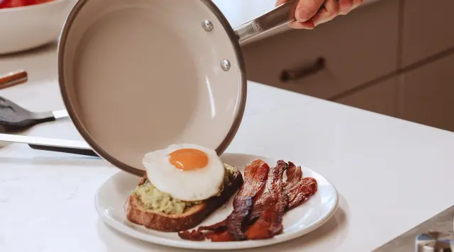 A hand is placing a frying pan over a plate of avocado toast topped with a fried egg and crispy bacon.