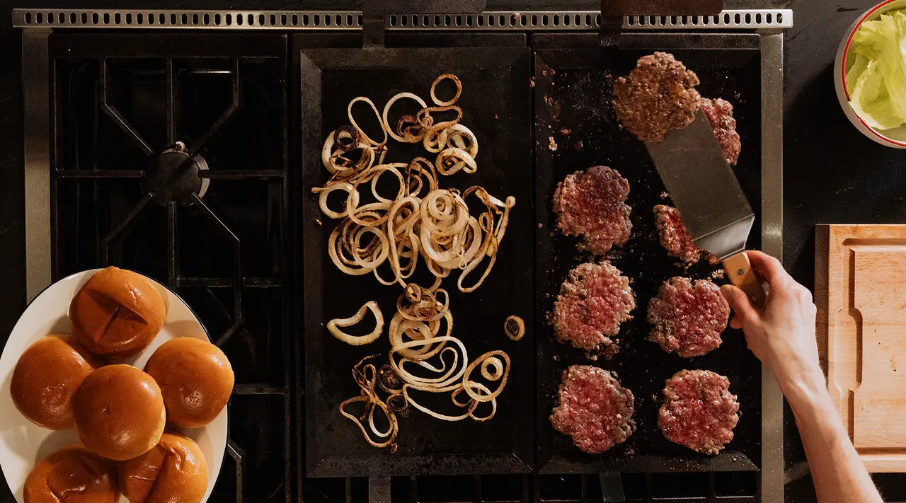 A person is cooking burgers and onions on a griddle, with a plate of buns nearby, suggesting preparation for a meal.