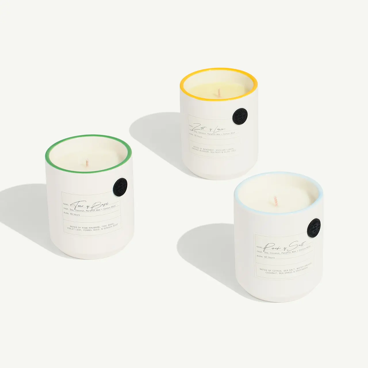 Three scented candles with different colored wax sit in a line casting soft shadows on a light background.