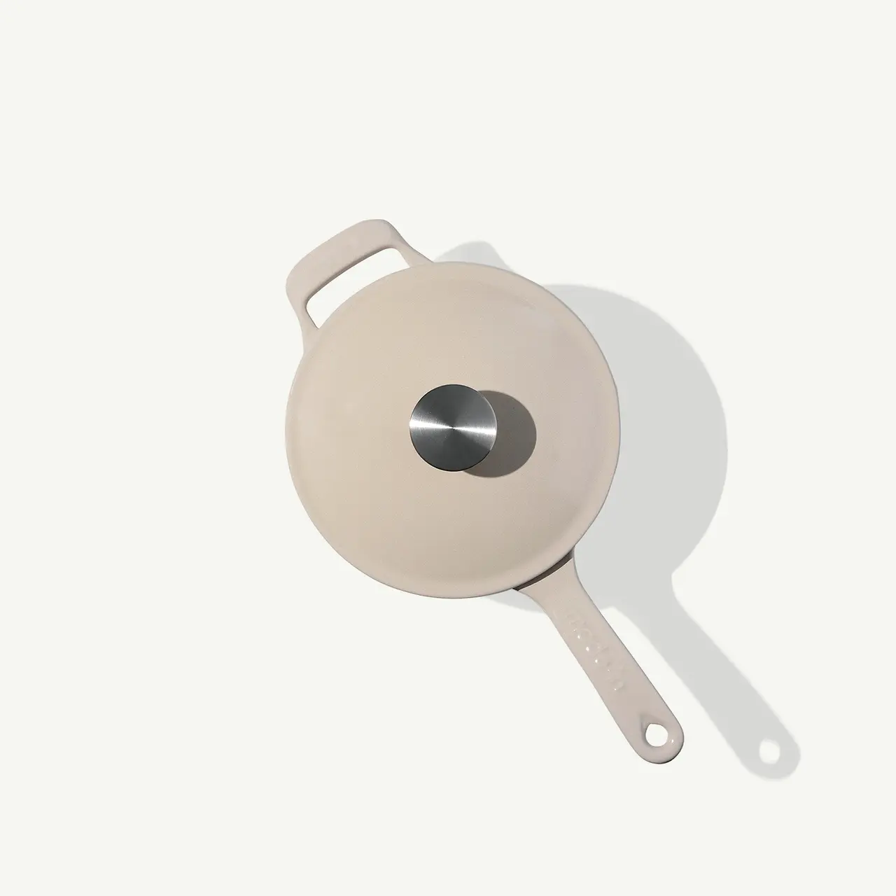 A beige-colored frying pan with a silver circular lid and an off-white handle casts a shadow on a light background.