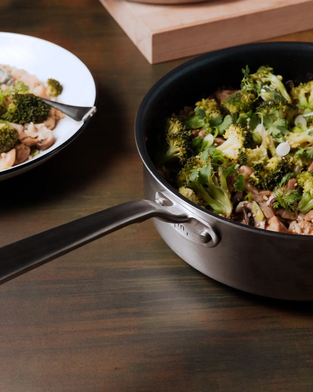 A skillet on a wooden table is filled with a stir-fried meal including broccoli and mushrooms, with a partially seen plate of the same meal in the background.