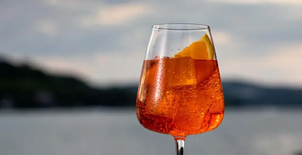 A glass of amber-colored beverage with ice and a slice of orange set against a blurred background of a water body under an overcast sky.