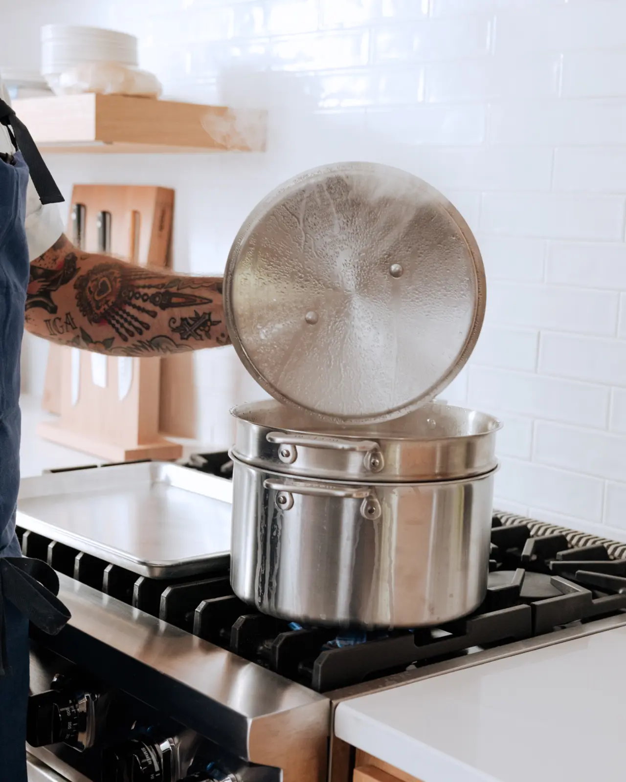 A person with a tattooed arm lifts the lid off a steaming pot on a stove.