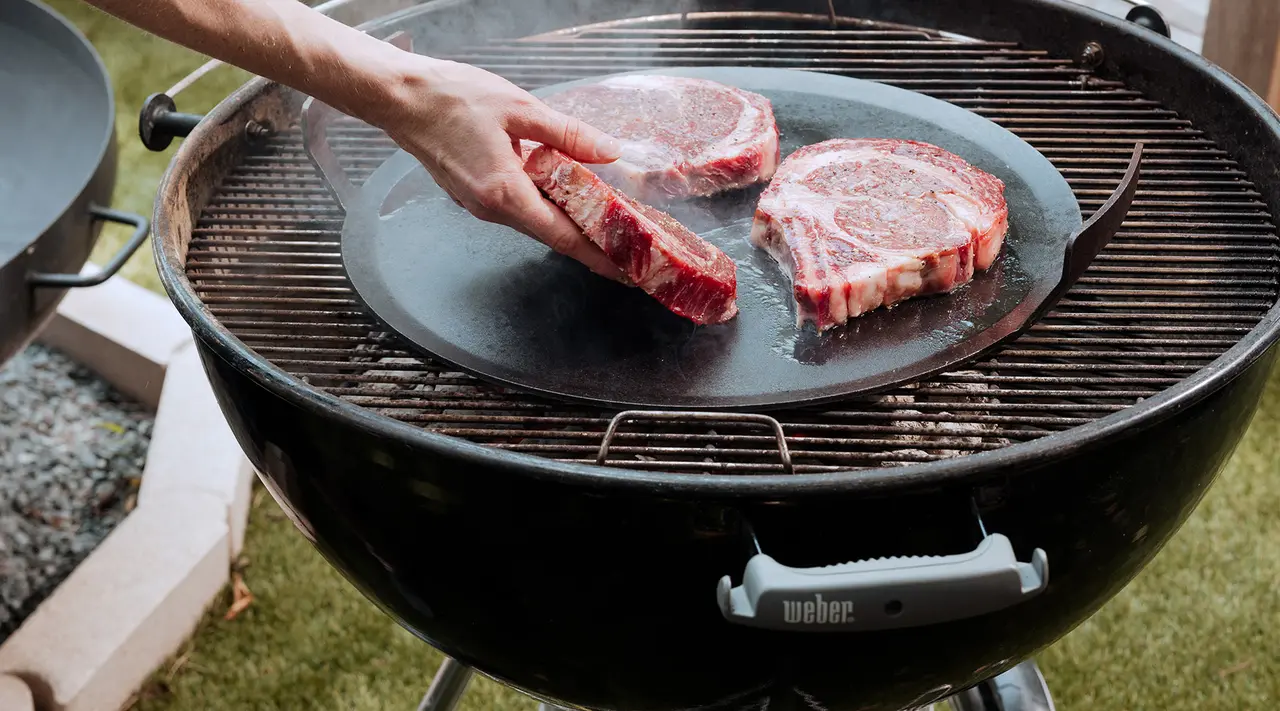 A hand places steaks on a smoking barbecue grill for cooking.