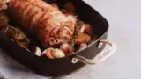 A golden-brown rolled roast sits atop a bed of seasoned potatoes and herbs in a black roasting pan.