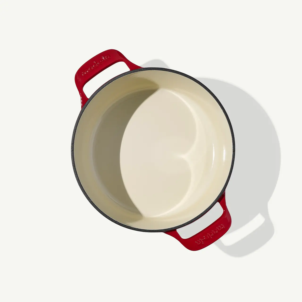 A top-down view of a cream-colored, empty Dutch oven with red handles on a light background.