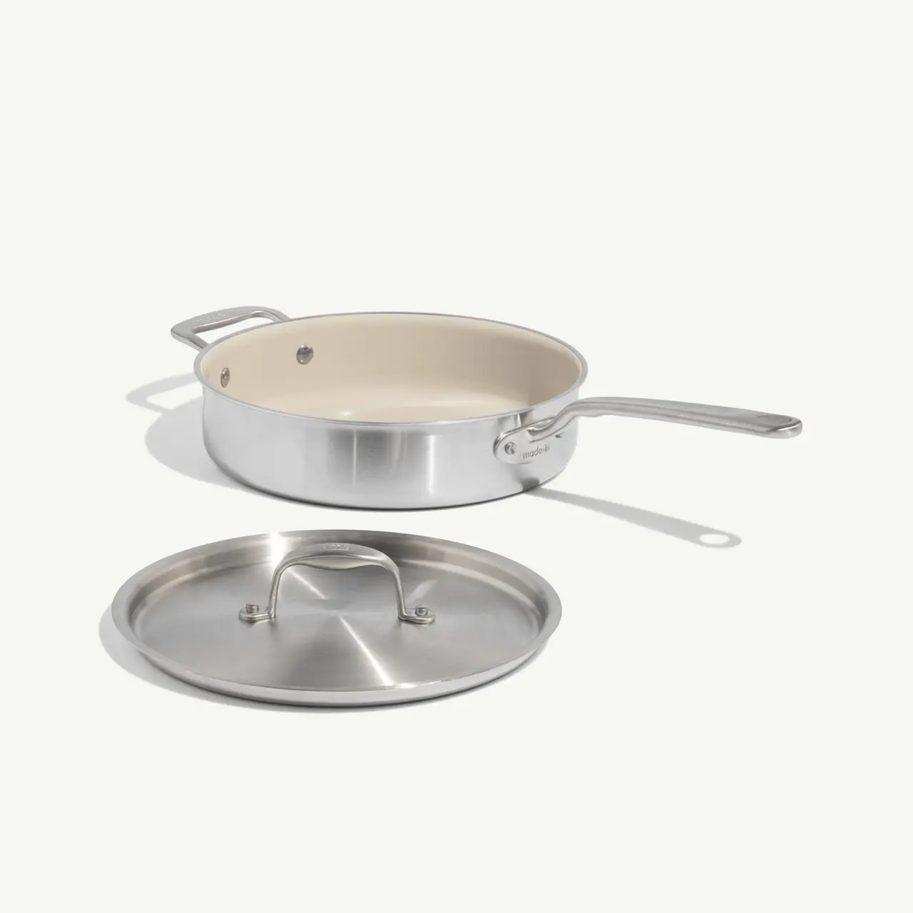 A stainless steel sauté pan with a white interior and a matching lid is displayed on a light background.