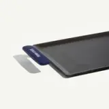 A rectangular, black baking sheet with a blue silicone handle on one side is displayed against a light background.