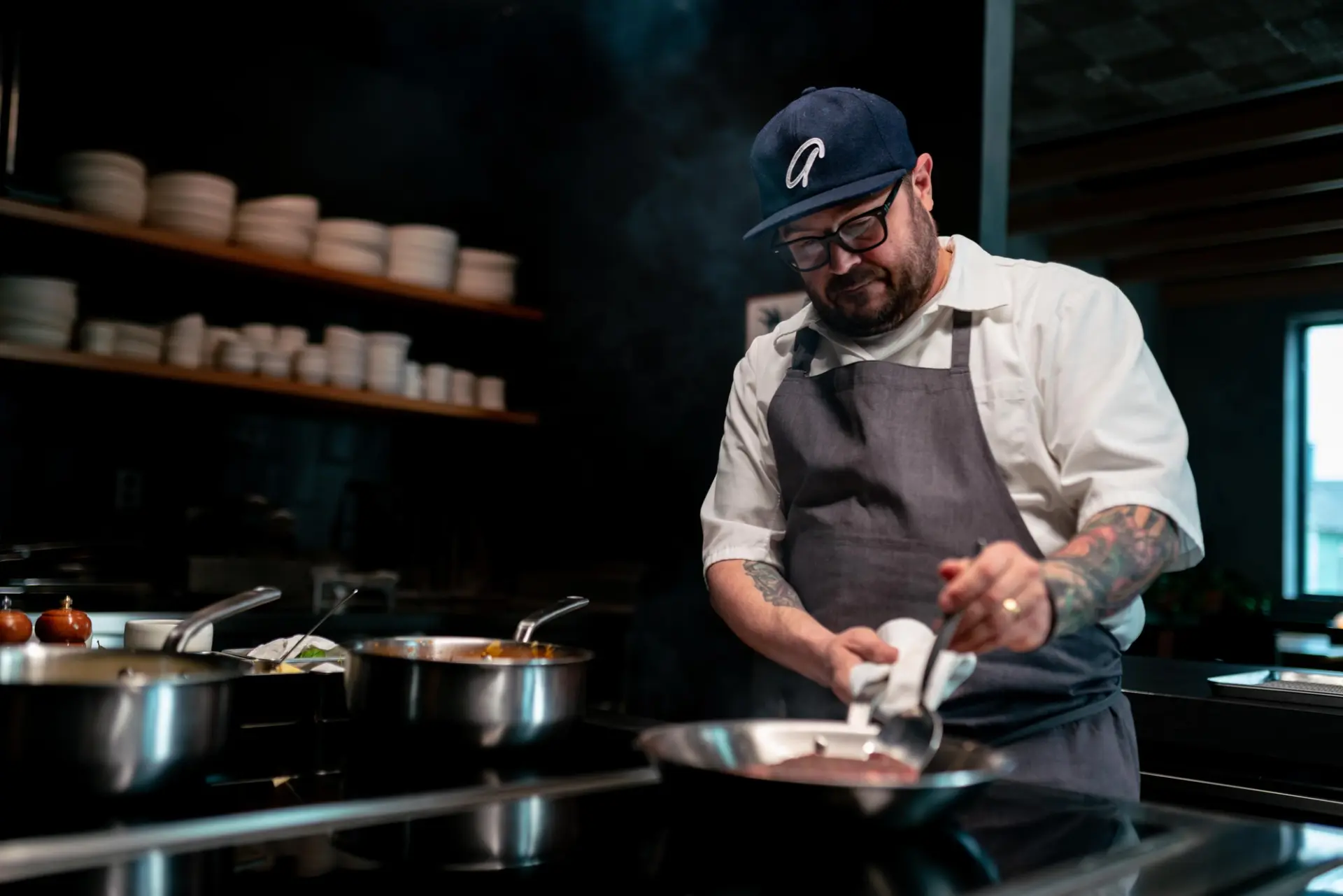 A chef in a gray apron and a baseball cap carefully prepares food on a stovetop, surrounded by cooking pots and a warm kitchen environment.