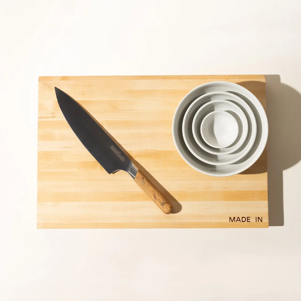 A chef's knife and a stack of white bowls are neatly placed on a bamboo cutting board under bright lighting.