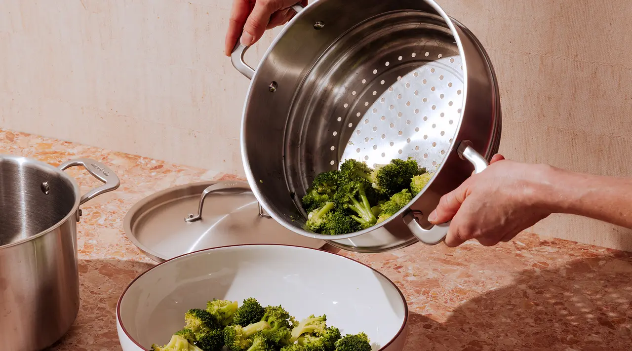 Hands are shown straining boiled broccoli into a bowl using a colander over a kitchen counter.