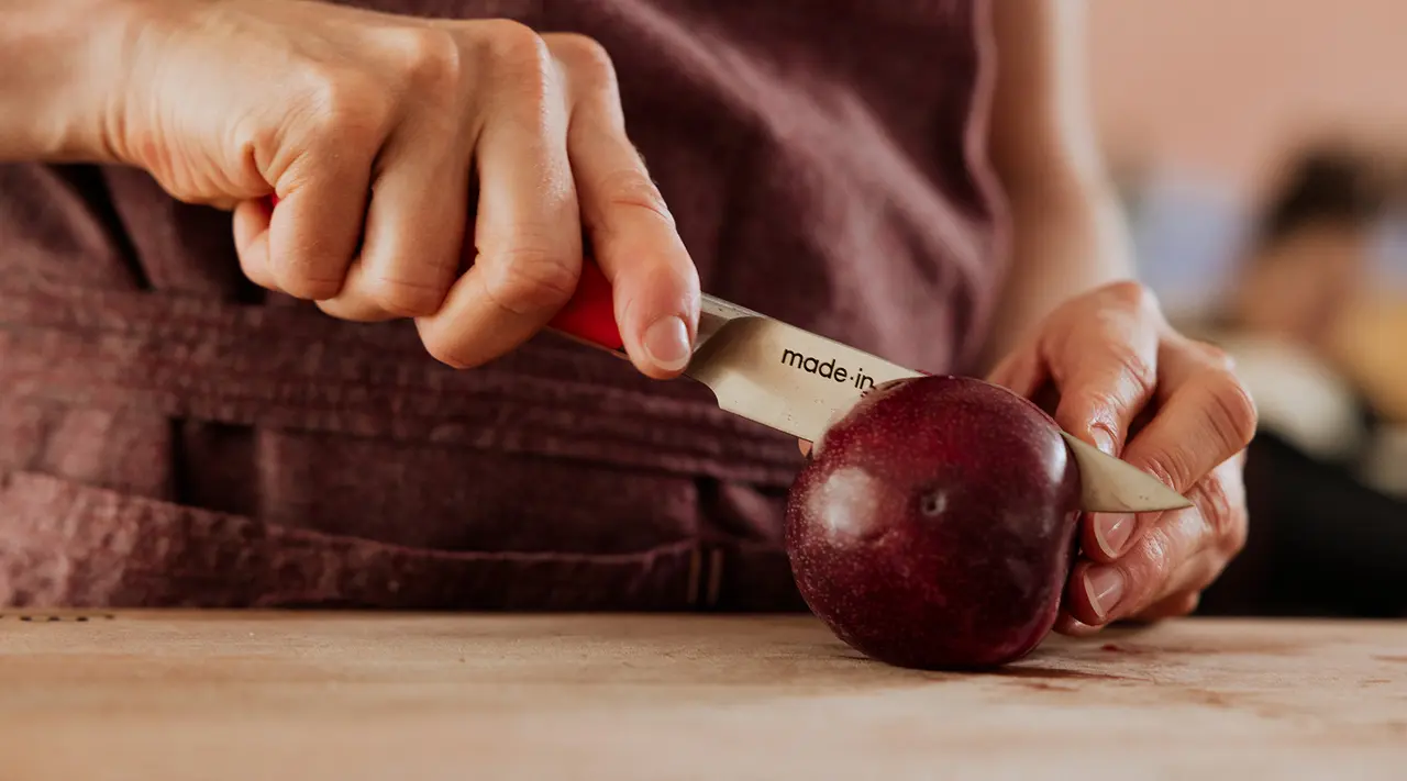 Hands are peeling a red apple with a vegetable peeler on a wooden surface.