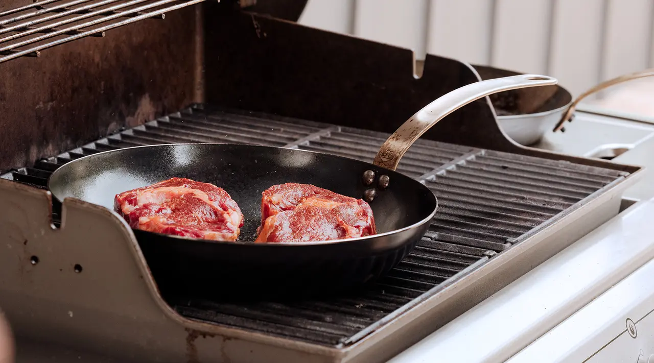 Two steaks are cooking in a frying pan on an outdoor grill.