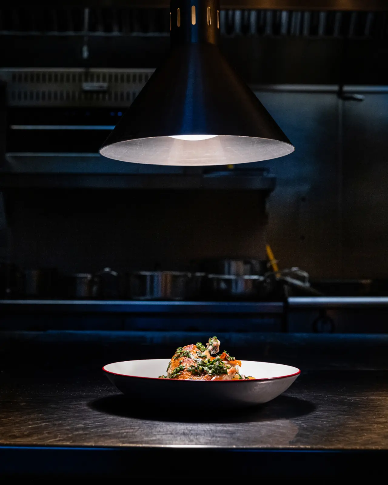 A spotlight illuminates a carefully plated dish on a counter in a dark professional kitchen setting.