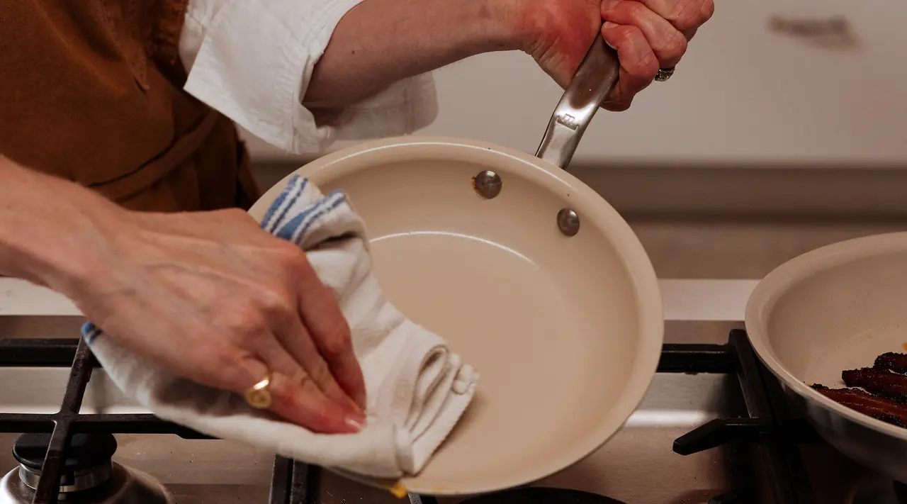 A person's hands are shown wiping a frying pan with a cloth over a stovetop.