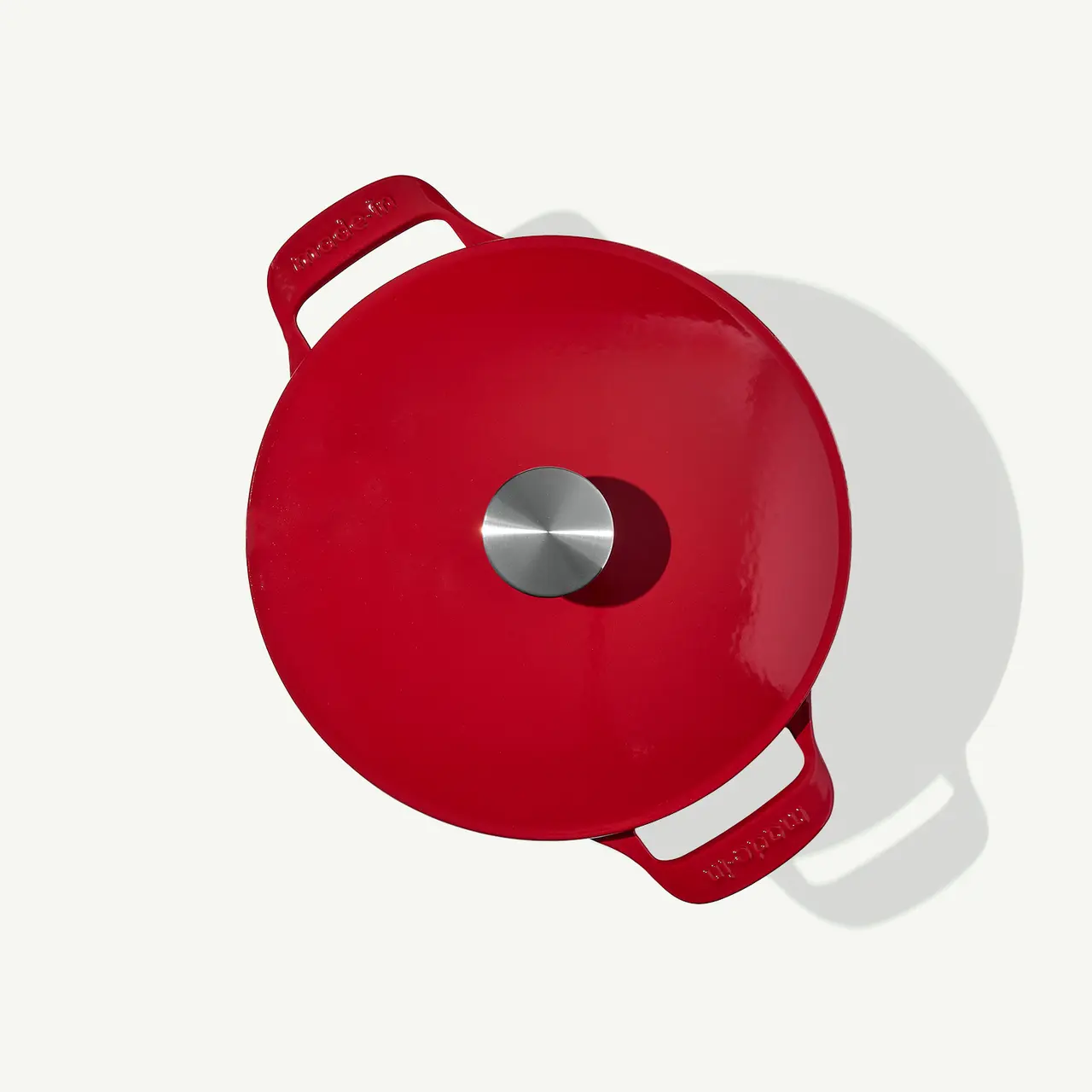 A red saucepan with two handles and a silver circular lid knob is viewed from above against a light background.