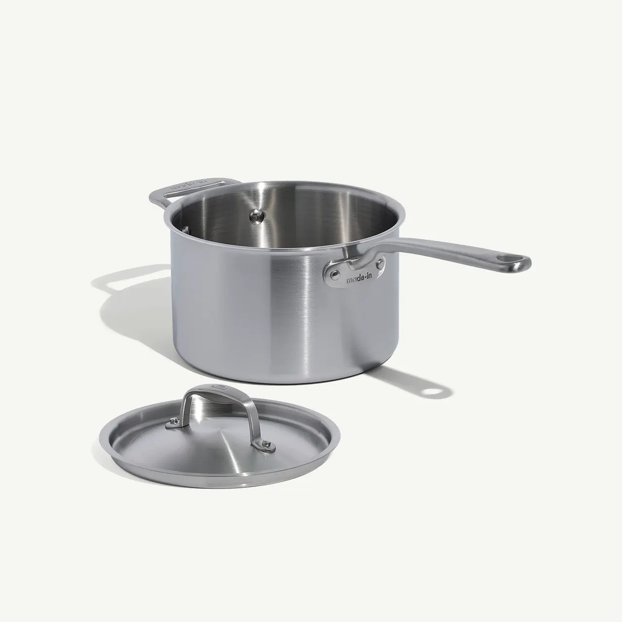 A stainless steel saucepan with a lid set to the side casts a soft shadow on a plain surface.