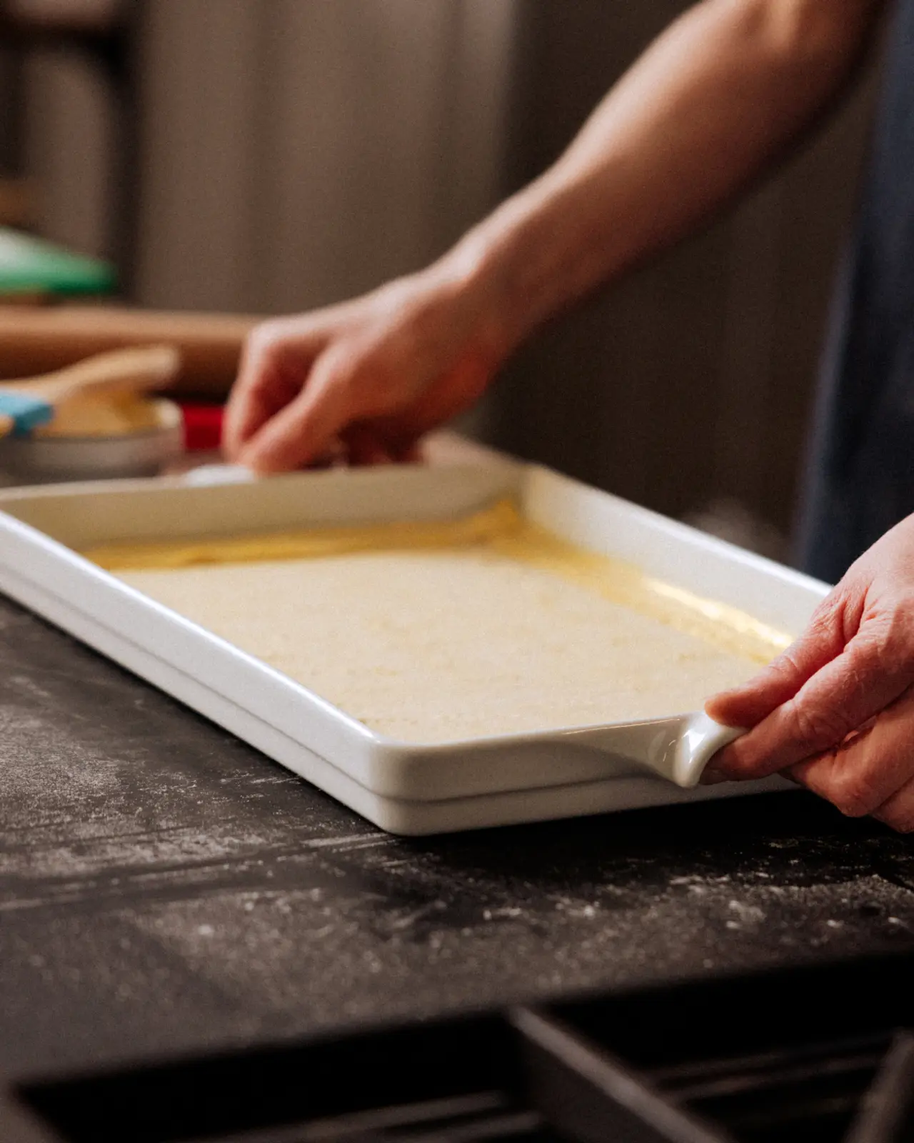 A person is smoothing batter in a white baking tray on a dark kitchen counter.