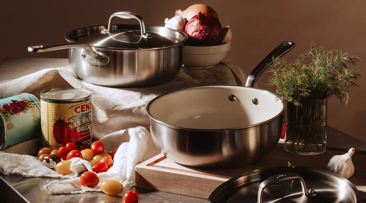 A variety of cooking utensils, including pots and pans, are arranged on a kitchen countertop alongside fresh vegetables and a can of tomatoes.