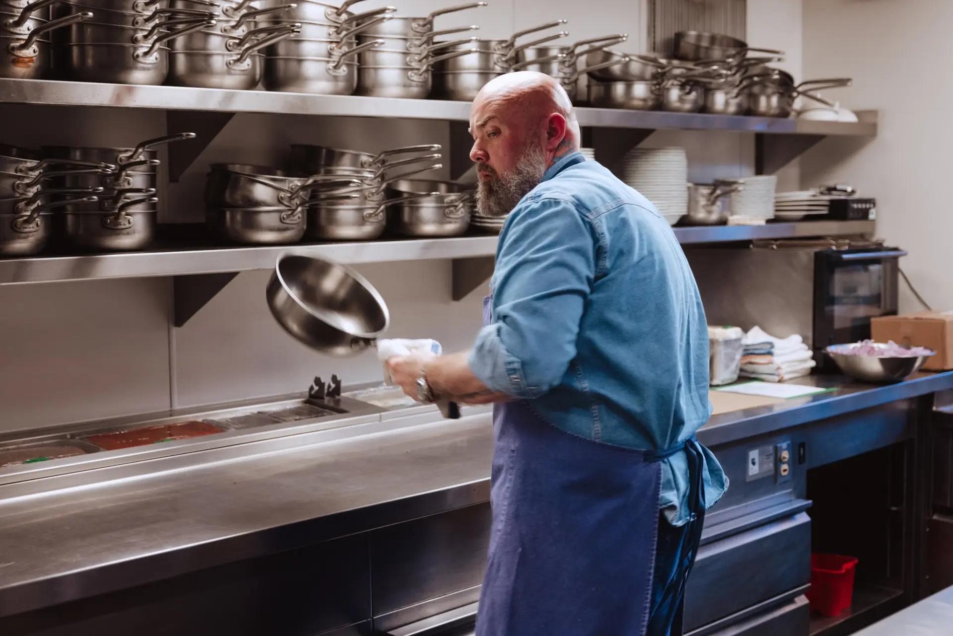 A chef in a blue shirt and apron expertly flips a skillet in a bustling kitchen filled with stainless steel cookware.