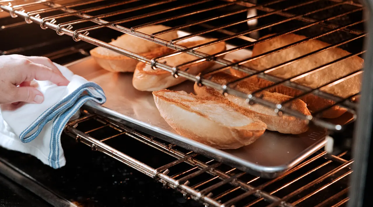 A person pulls out a tray of freshly baked baguettes from an oven using a cloth to protect their hand.