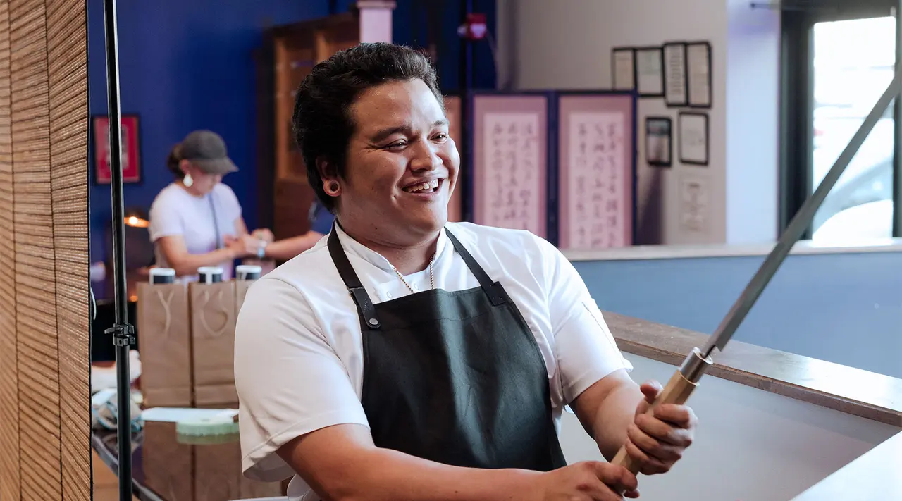 A smiling chef wearing a white shirt and apron holds a long sword or knife as if playfully wielding it, standing in a restaurant kitchen with another person working in the background.