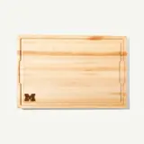 A wooden cutting board with a carved letter "M" in one corner.