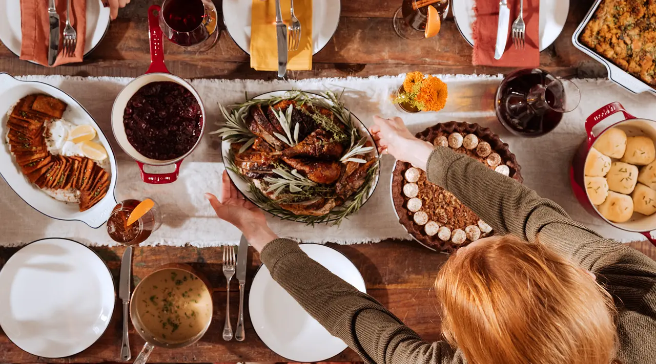 A traditional Thanksgiving meal with various dishes displayed on a table and a person reaching out to the roasted turkey centerpiece.