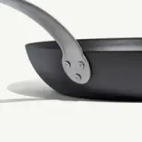 A close-up of a black frying pan showing the connection of the handle to the pan with three rivets on a light background.