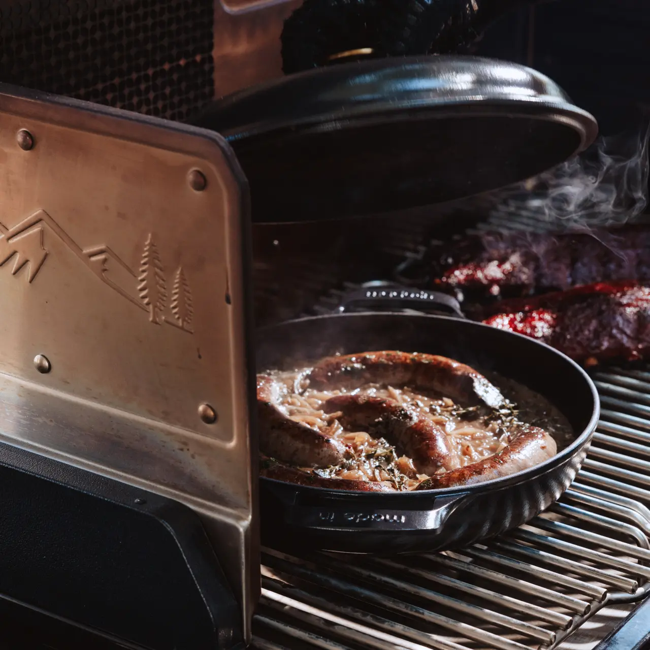 Sizzling sausages cook in a skillet on a grill with steam rising, and another meat is visible in the background.