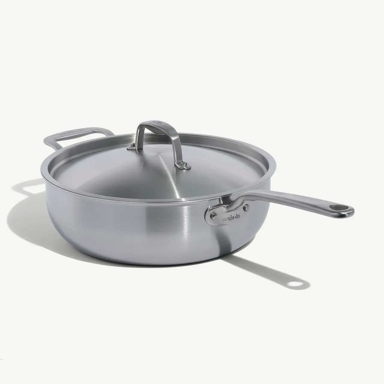 A stainless steel sauté pan with a lid sits on a neutral background, reflecting a light source.