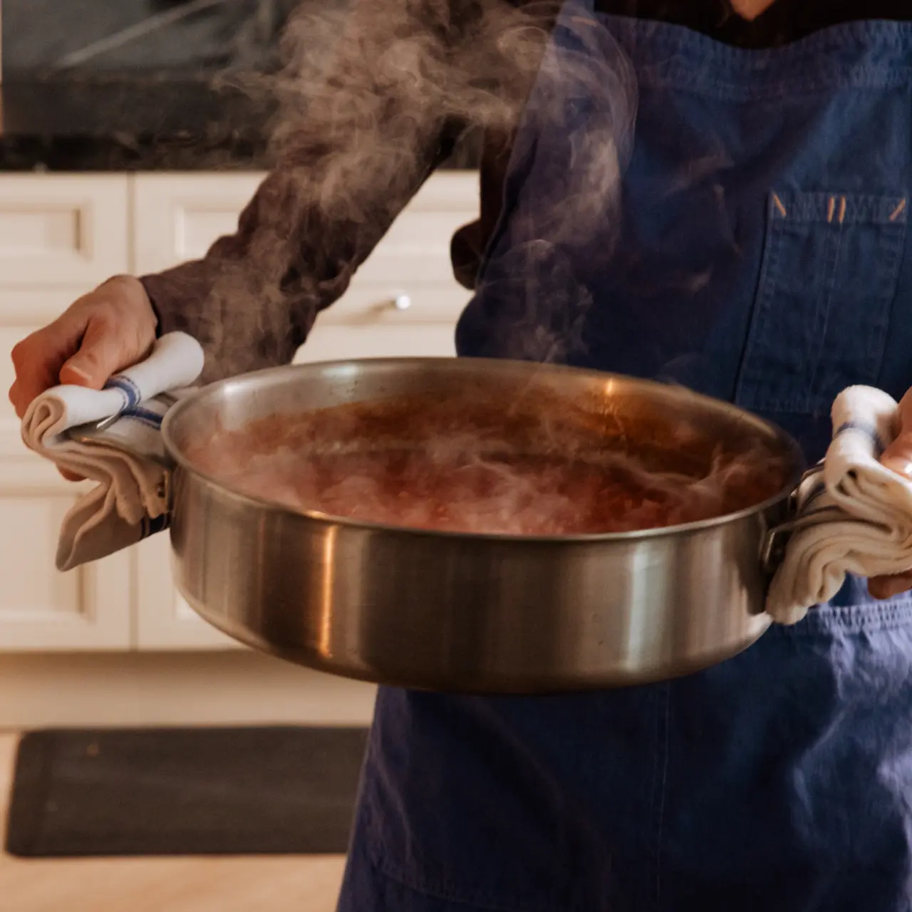 A person wearing an apron holds a steaming pot with both hands using oven mitts.
