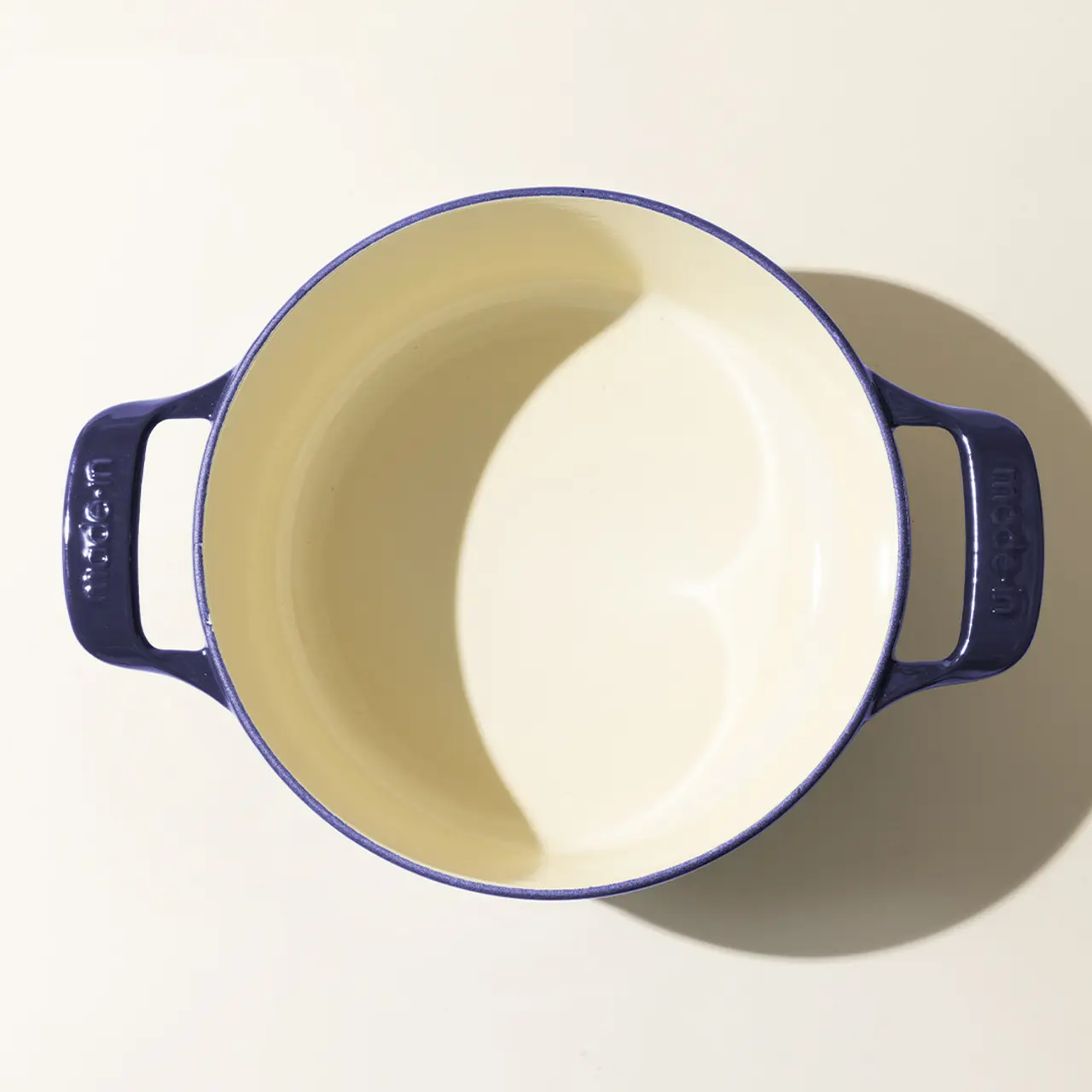Top-down view of an empty, cream-colored round casserole dish with two navy blue handles, casting a shadow on a light surface.