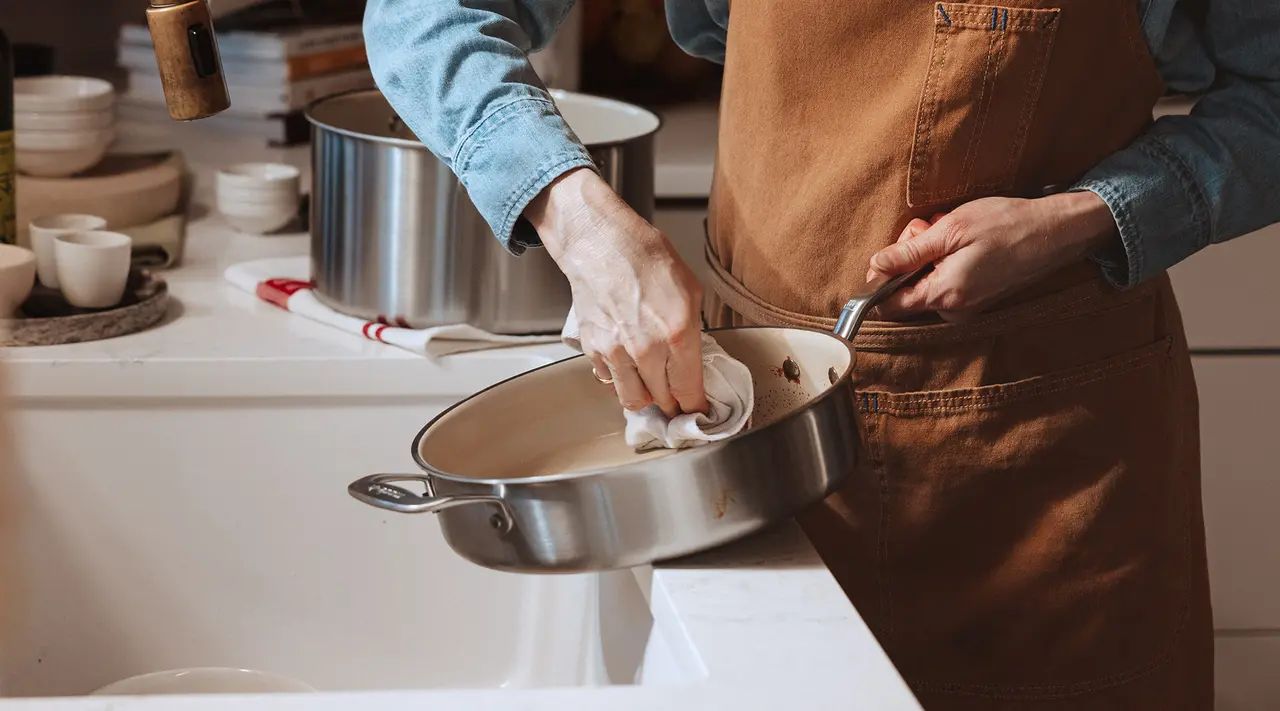 A person wearing an apron is cleaning a frying pan in a kitchen sink.