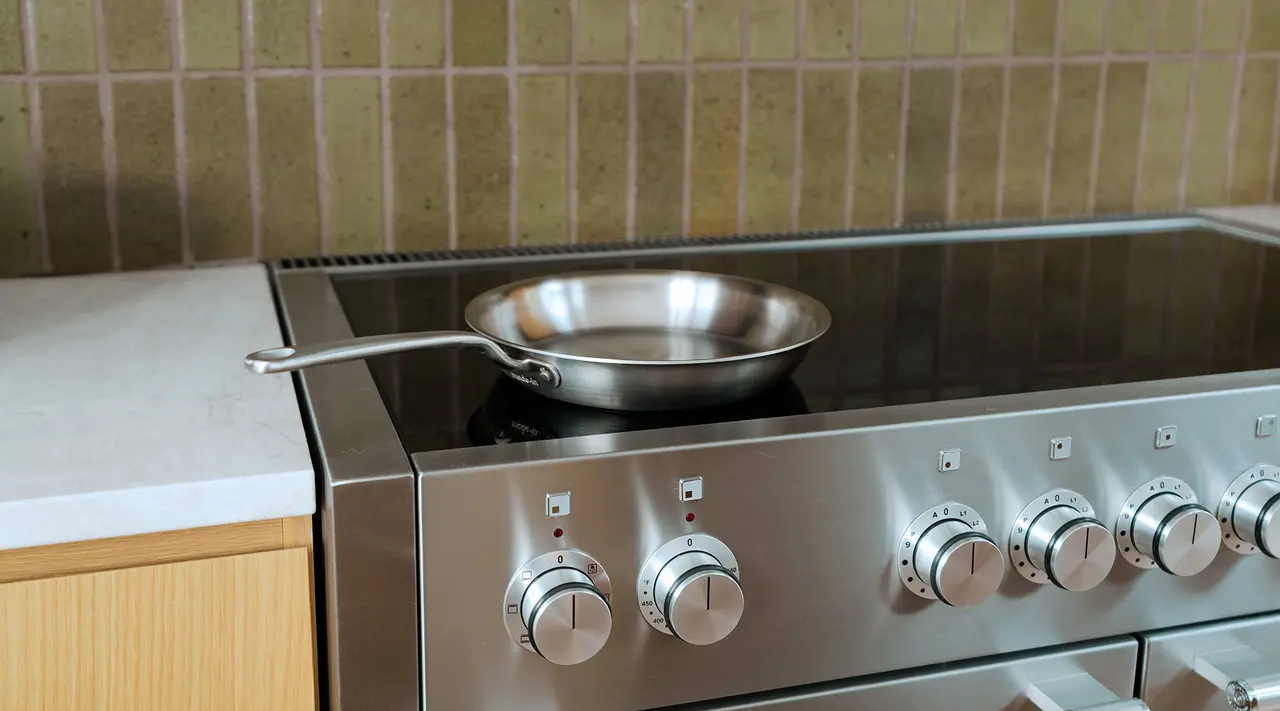 A stainless steel frying pan sits on a modern stove with sleek control knobs against a tiled backsplash.