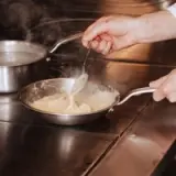 A hand holds a spoon, stirring a creamy substance in a pan on a stovetop, with steam rising in the background.