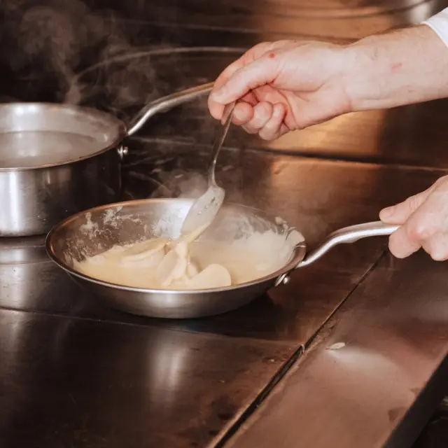A hand holds a spoon, stirring a creamy substance in a pan on a stovetop, with steam rising in the background.