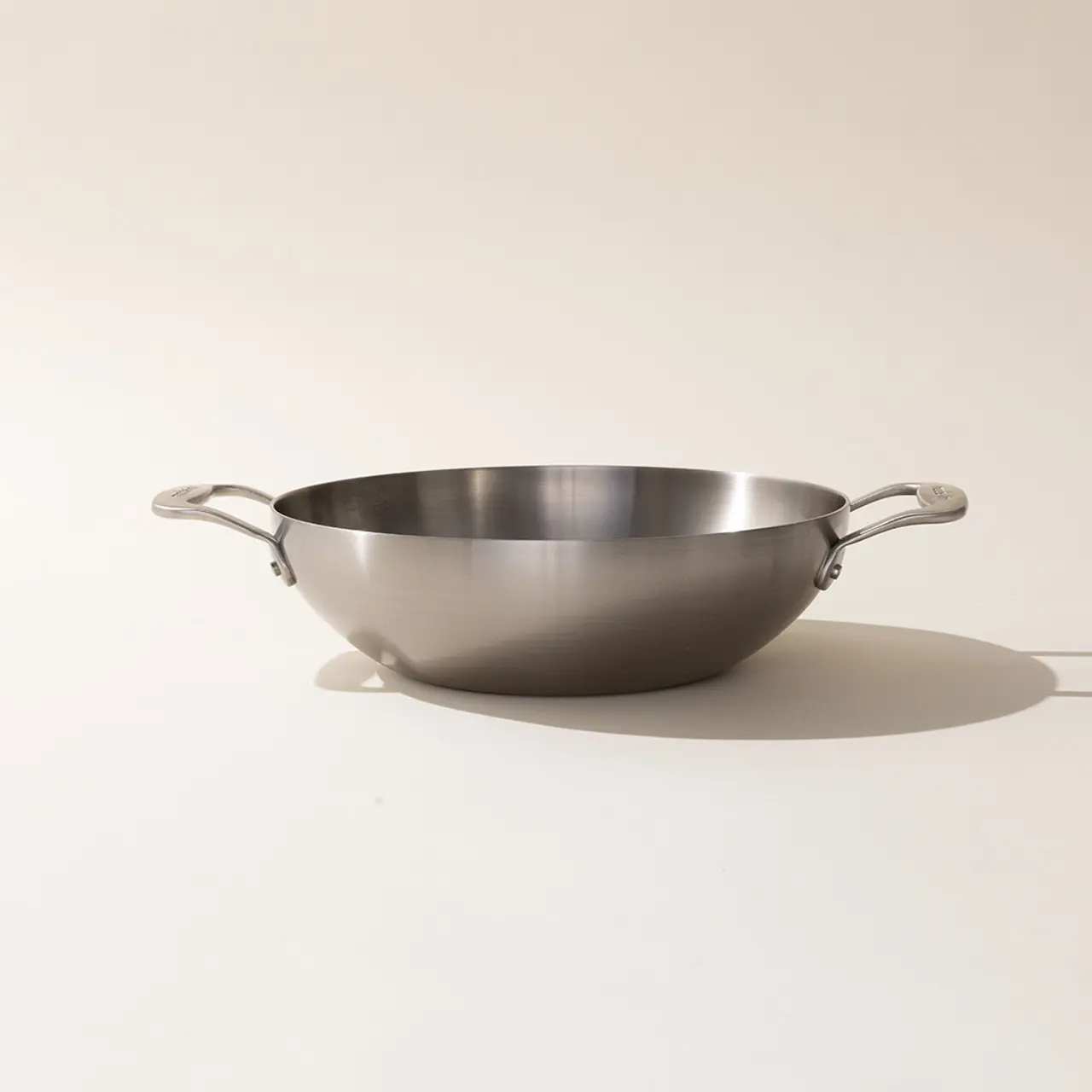 A stainless steel wok with two handles against a neutral background.