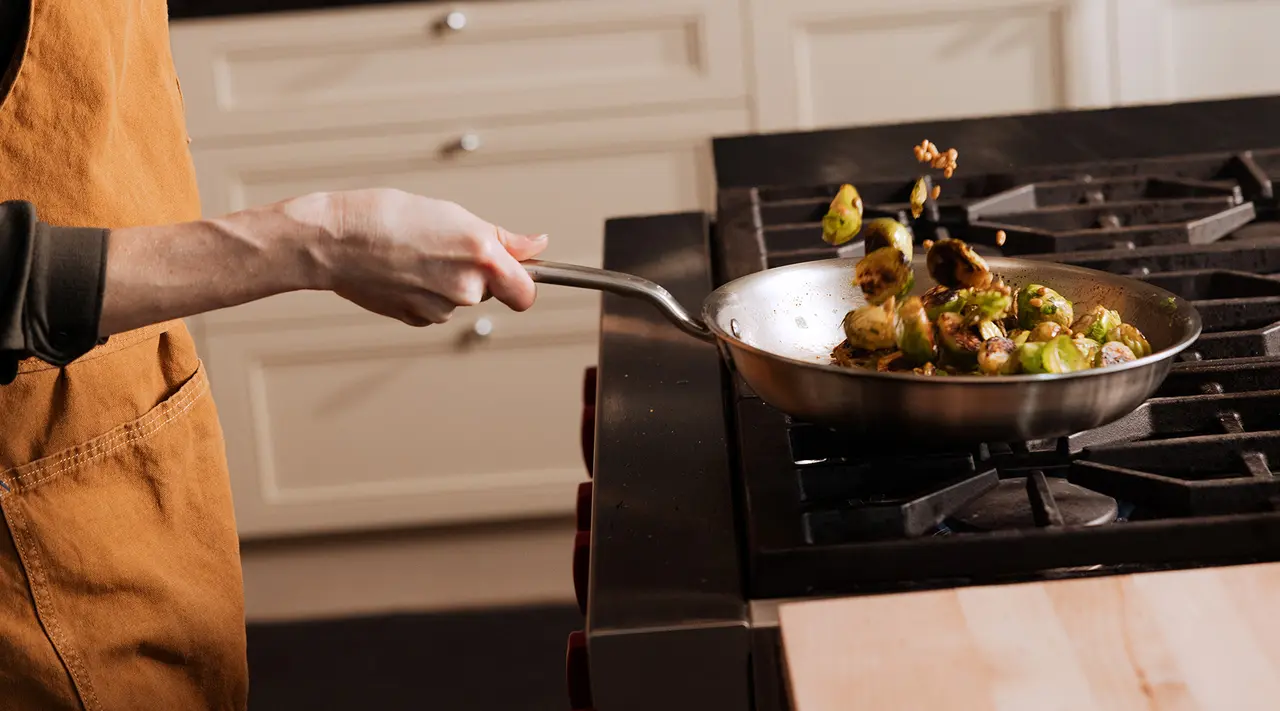 A person in an apron flips Brussels sprouts in a sauté pan over a stove.