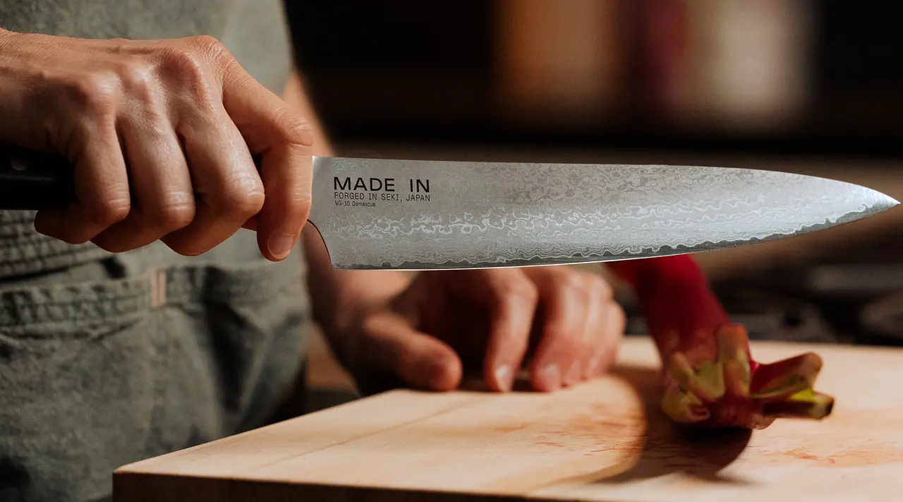 A person is holding a chef's knife with "MADE IN" text visible on it, hovering over a cutting board where some vegetable cutting seems to be in progress.