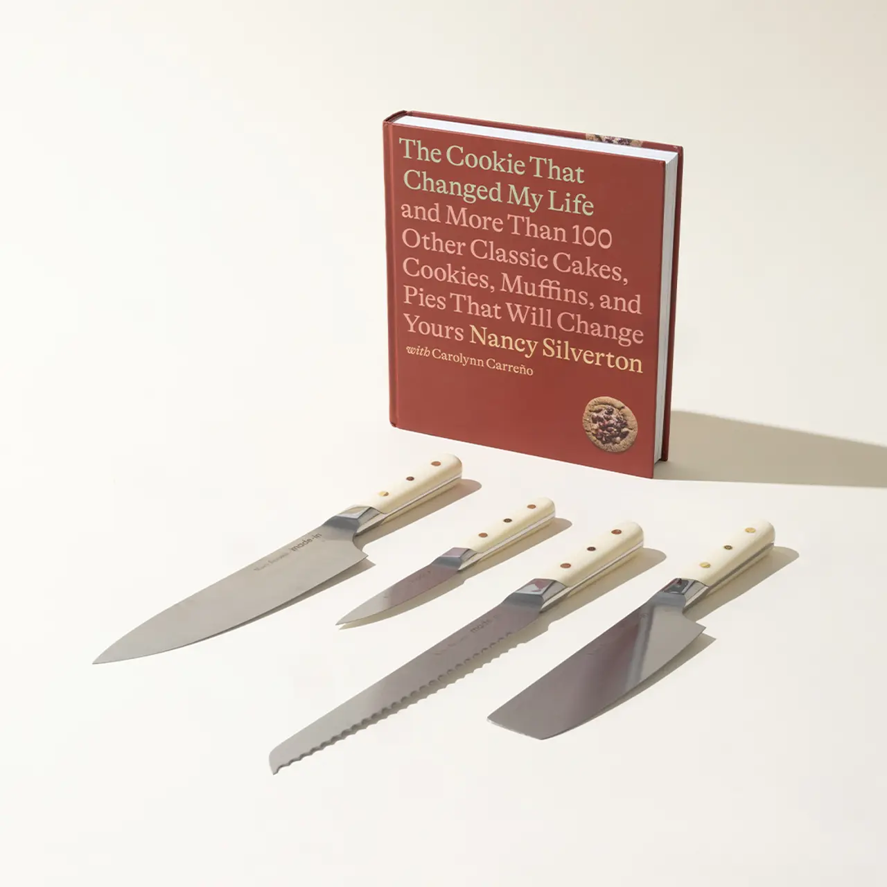 A cookbook titled "The Cookie That Changed My Life" stands behind a neatly arranged row of four kitchen knives on a light background.