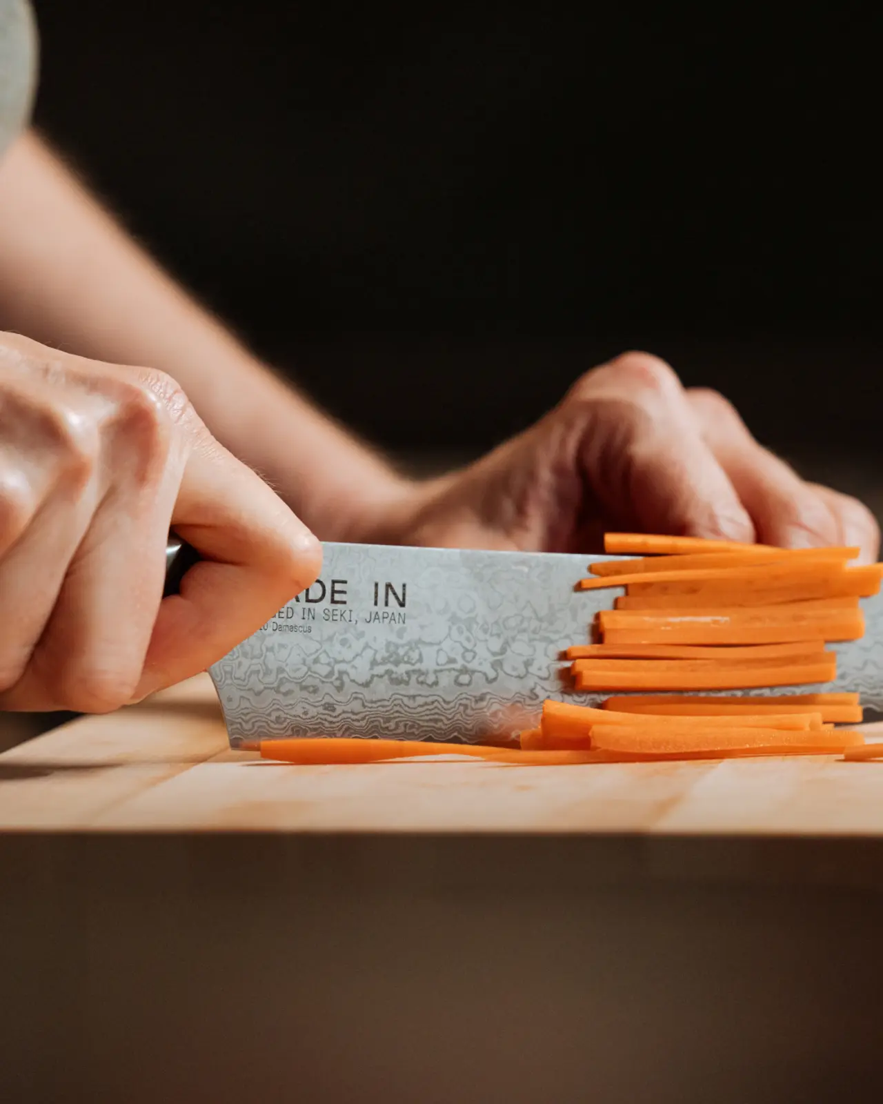 Hands are slicing carrots on a wooden cutting board with a steel kitchen knife.