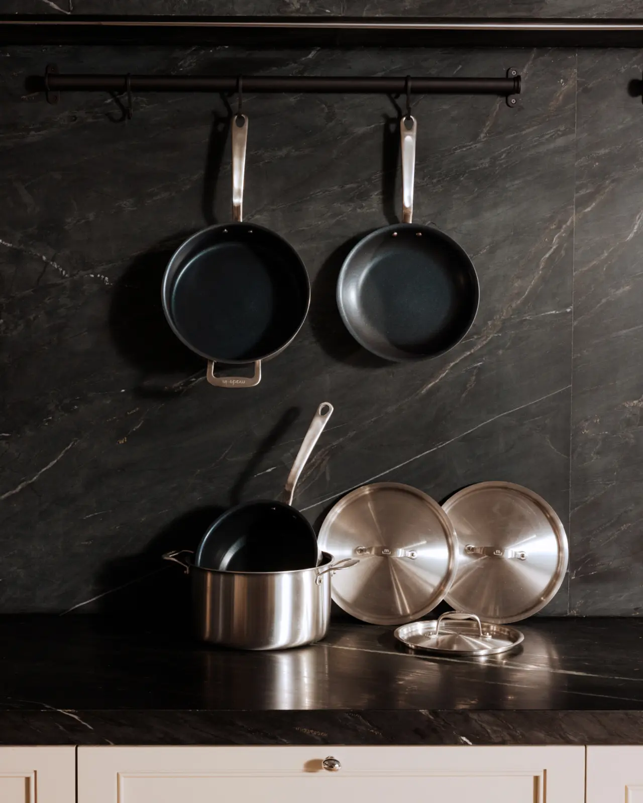 Hanging pans and pots with lids rest on a kitchen counter against a dark marble backsplash.