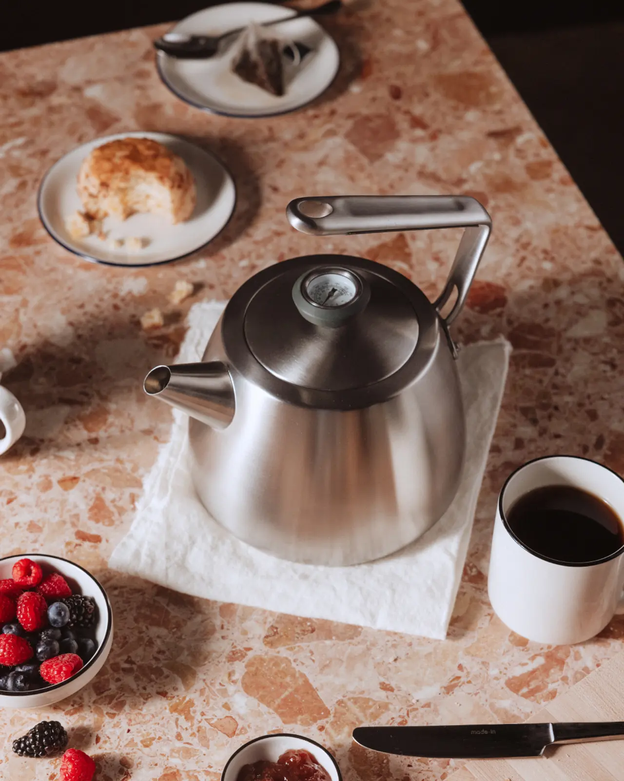 A stainless steel kettle sits on a white napkin, surrounded by a cup of coffee, a plate of cake, and bowls of mixed berries and jam on a marble table.