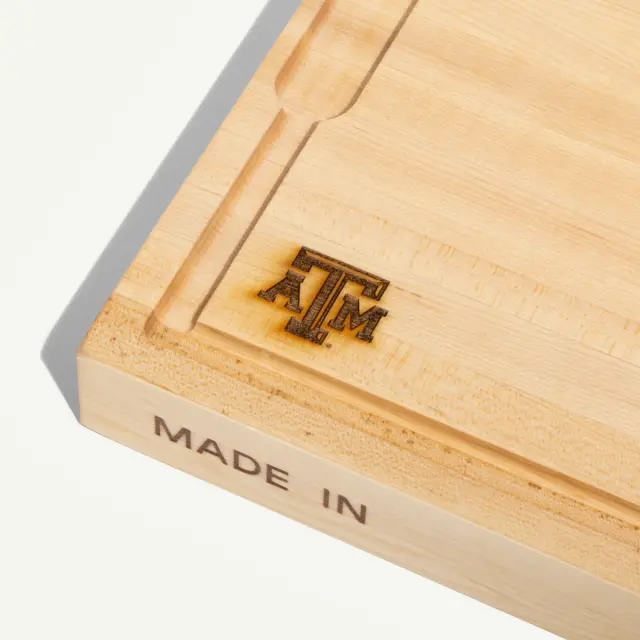 A wooden cutting board features the engraved letters "TAMU" and the phrase "MADE IN" on its side.