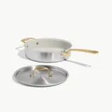 A silver sauté pan with a white interior and a gold handle is positioned next to its matching lid.