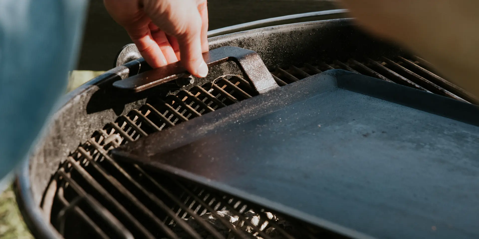 A person's hand is shown lifting the lid of a barbecue grill, revealing a grill grate inside.