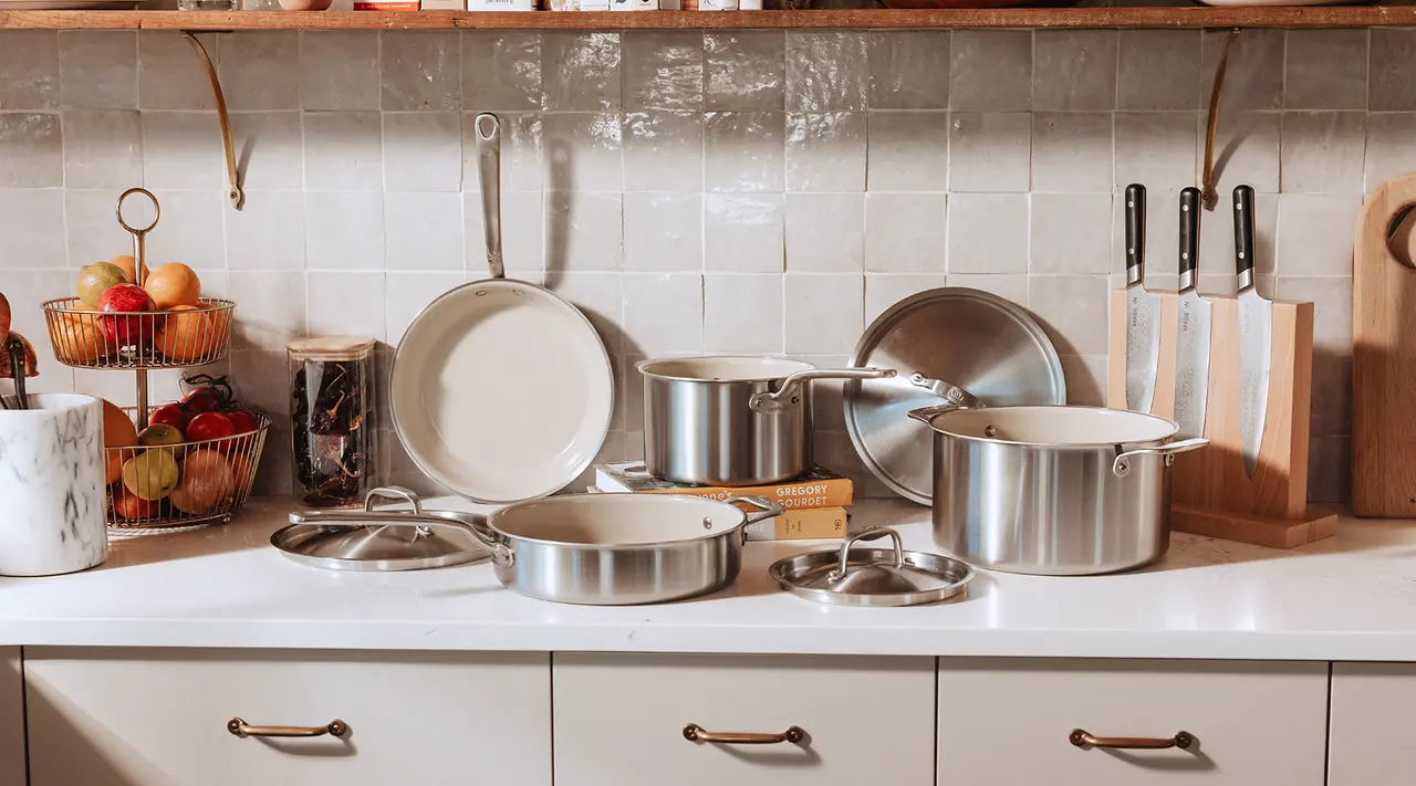 A clean kitchen countertop displays various stainless steel pots and pans alongside a fruit basket and knife block.