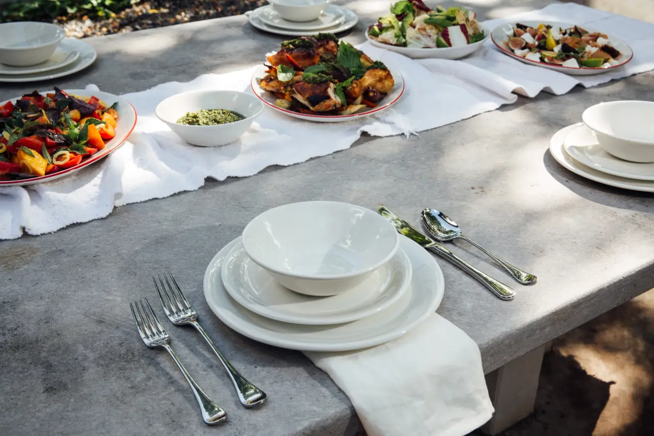 A table is set outdoors with stacked white plates, silverware, and multiple dishes of food ready for a meal.